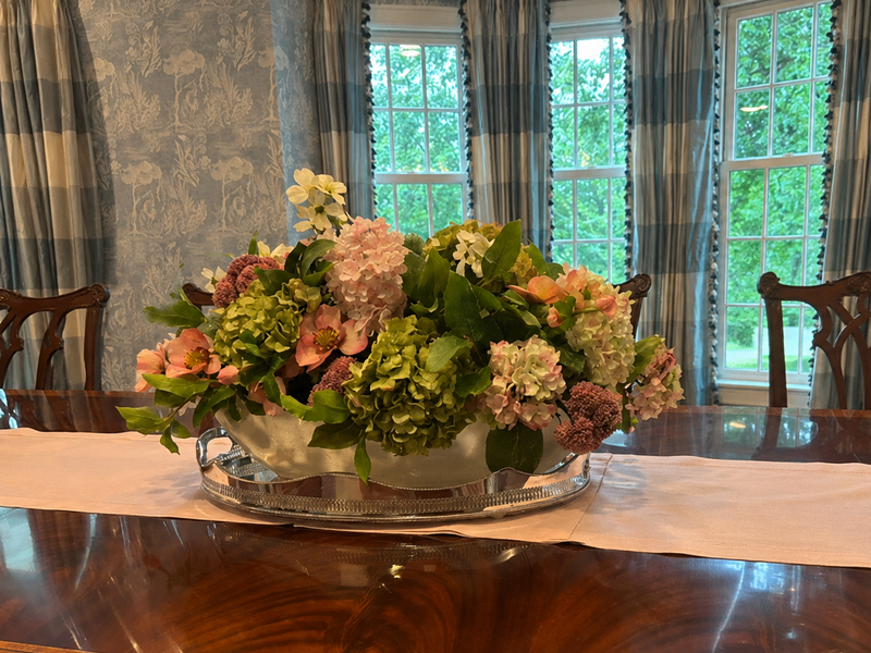 Floral arrangement on a wooden dining table with large windows and curtains in the background