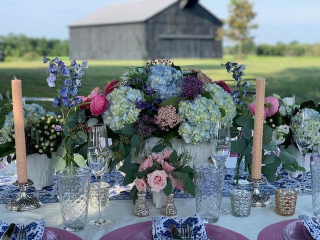 Decorative table setting with flowers, candles, and glasses outdoors near a barn.