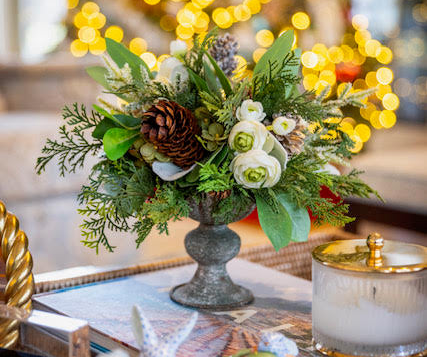 Decorative floral arrangement with greenery and pinecones on a table with blurred lights in the background